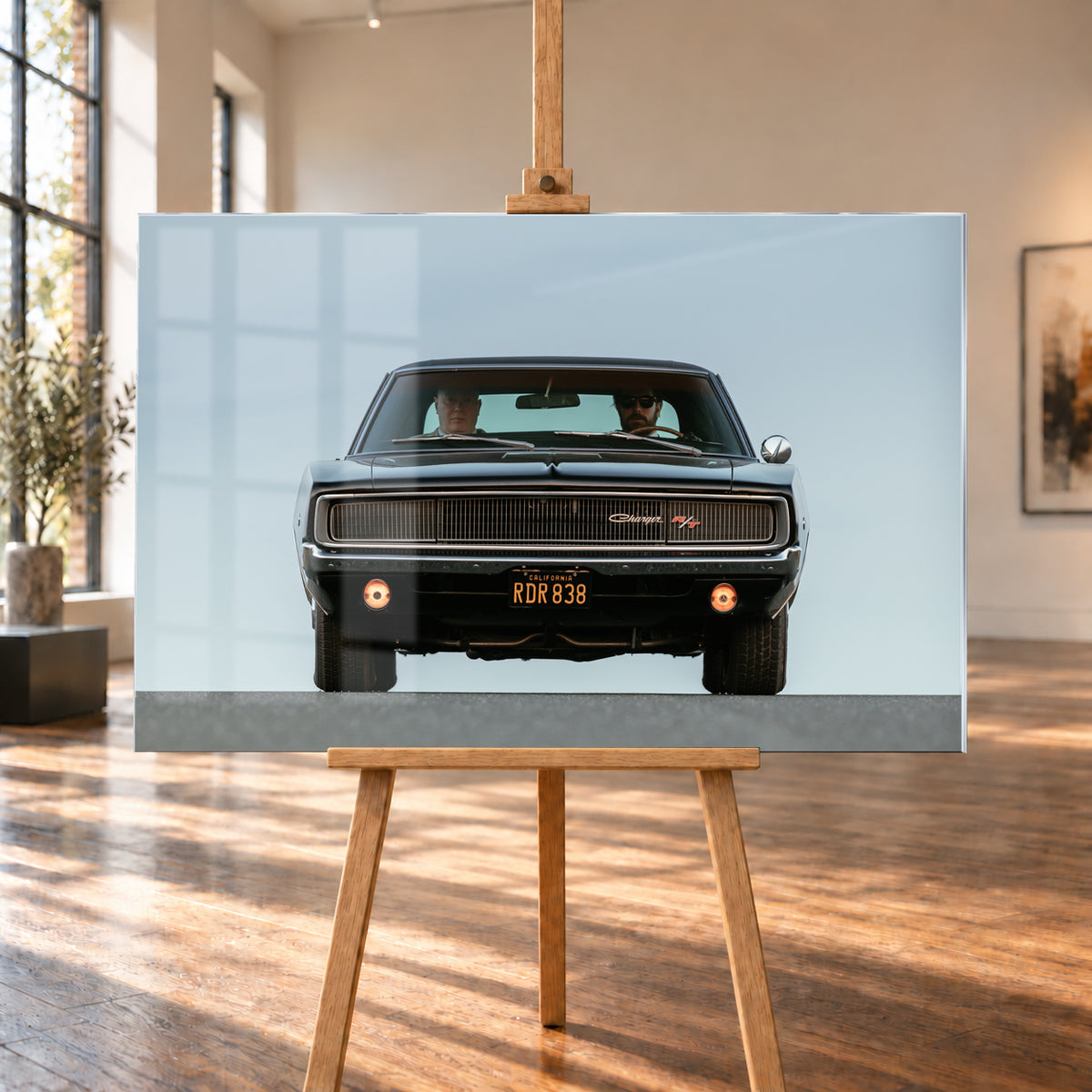 Sehr dunkler Dodge Charger R/T in tiefer, zentraler Frontalansicht vor blassblauem Himmel; zwei Personen sitzen hinter der Windschutzscheibe, zwei Zusatzleuchten glimmen warm; Visualisierung als Acrylglas-Wandbild, auf einer Holzstaffelei in einem hellen Galerieraum.