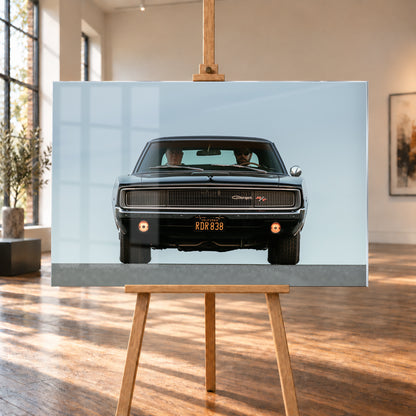 Sehr dunkler Dodge Charger R/T in tiefer, zentraler Frontalansicht vor blassblauem Himmel; zwei Personen sitzen hinter der Windschutzscheibe, zwei Zusatzleuchten glimmen warm; Visualisierung als Acrylglas-Wandbild, auf einer Holzstaffelei in einem hellen Galerieraum.