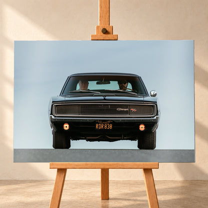 Sehr dunkler Dodge Charger R/T in tiefer, zentraler Frontalansicht vor blassblauem Himmel; zwei Personen sitzen hinter der Windschutzscheibe, zwei Zusatzleuchten glimmen warm; Visualisierung als Alu gebürstet-Wandbild, frontal auf einer Holzstaffelei.