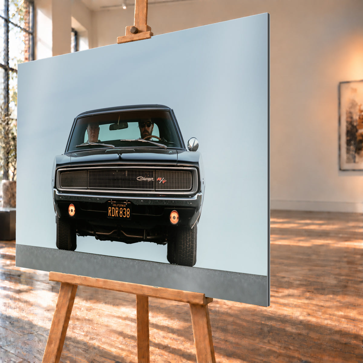 Sehr dunkler Dodge Charger R/T in tiefer, zentraler Frontalansicht vor blassblauem Himmel; zwei Personen sitzen hinter der Windschutzscheibe, zwei Zusatzleuchten glimmen warm; Visualisierung als Alu gebürstet-Wandbild, schräg von der Seite auf einer Holzstaffelei.