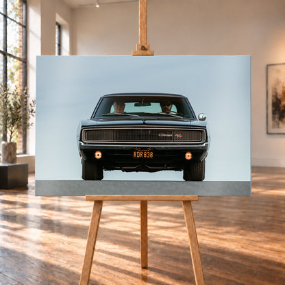 Sehr dunkler Dodge Charger R/T in tiefer, zentraler Frontalansicht vor blassblauem Himmel; zwei Personen sitzen hinter der Windschutzscheibe, zwei Zusatzleuchten glimmen warm; Visualisierung als Alu gebürstet-Wandbild, auf einer Holzstaffelei in einem hellen Galerieraum.