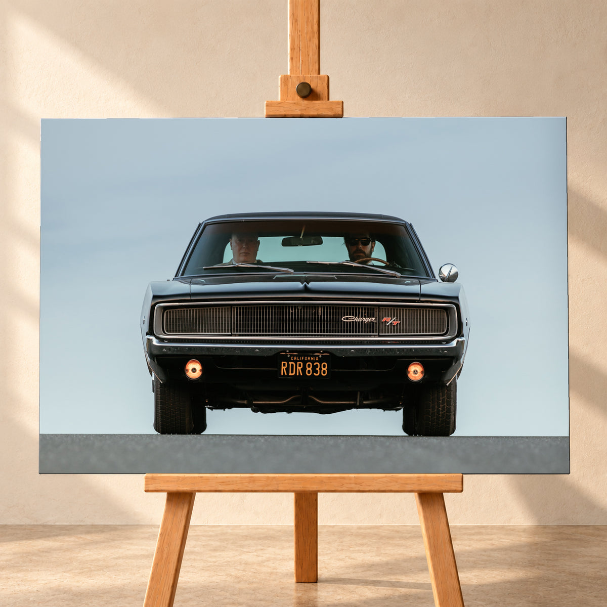 Sehr dunkler Dodge Charger R/T in tiefer, zentraler Frontalansicht vor blassblauem Himmel; zwei Personen sitzen hinter der Windschutzscheibe, zwei Zusatzleuchten glimmen warm; Visualisierung als Leinwand-Wandbild, frontal auf einer Holzstaffelei.