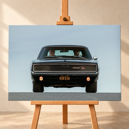 Sehr dunkler Dodge Charger R/T in tiefer, zentraler Frontalansicht vor blassblauem Himmel; zwei Personen sitzen hinter der Windschutzscheibe, zwei Zusatzleuchten glimmen warm; Visualisierung als Leinwand-Wandbild, frontal auf einer Holzstaffelei.