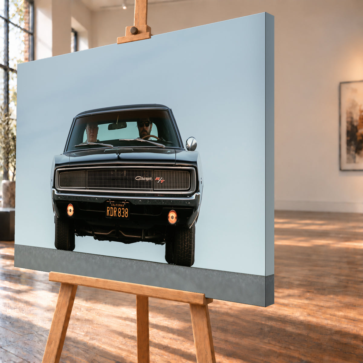 Sehr dunkler Dodge Charger R/T in tiefer, zentraler Frontalansicht vor blassblauem Himmel; zwei Personen sitzen hinter der Windschutzscheibe, zwei Zusatzleuchten glimmen warm; Visualisierung als Leinwand-Wandbild, schräg von der Seite auf einer Holzstaffelei.