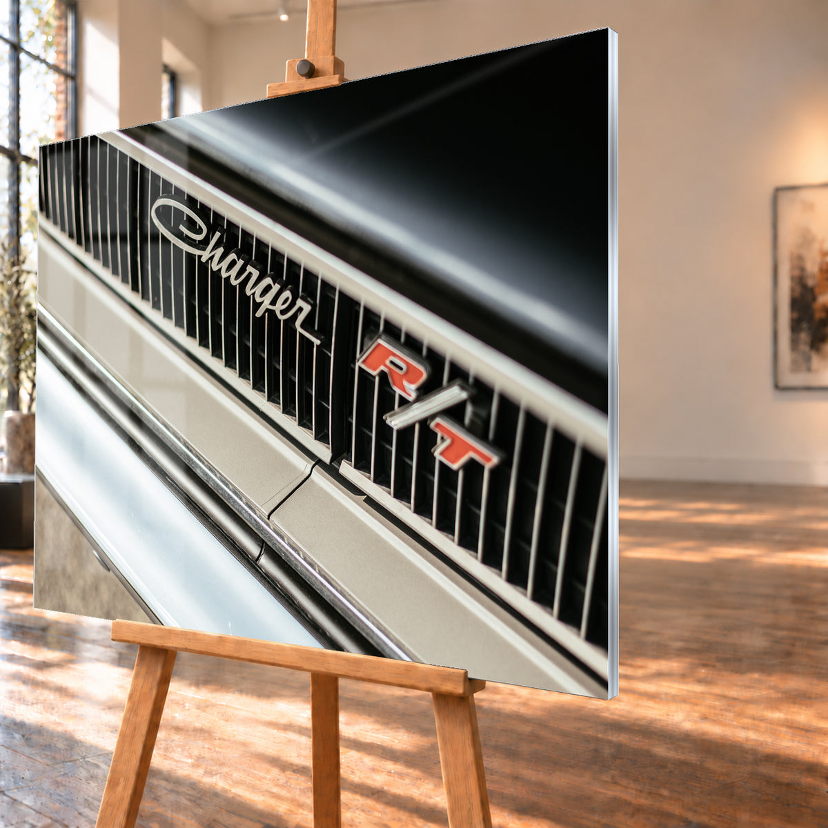 Diagonale Nahaufnahme eines hell lackierten Charger R/T mit schwarzem Lamellengrill, verchromtem Charger-Schriftzug und rot-weißem R/T-Emblem; Chromleisten reflektieren helles Licht; Visualisierung als Acrylglas-Wandbild, schräg von der Seite auf einer Holzstaffelei.