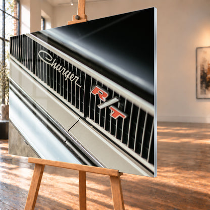 Diagonale Nahaufnahme eines hell lackierten Charger R/T mit schwarzem Lamellengrill, verchromtem Charger-Schriftzug und rot-weißem R/T-Emblem; Chromleisten reflektieren helles Licht; Visualisierung als Acrylglas-Wandbild, schräg von der Seite auf einer Holzstaffelei.