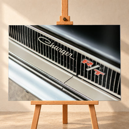 Diagonale Nahaufnahme eines hell lackierten Charger R/T mit schwarzem Lamellengrill, verchromtem Charger-Schriftzug und rot-weißem R/T-Emblem; Chromleisten reflektieren helles Licht; Visualisierung als Alu gebürstet-Wandbild, frontal auf einer Holzstaffelei.
