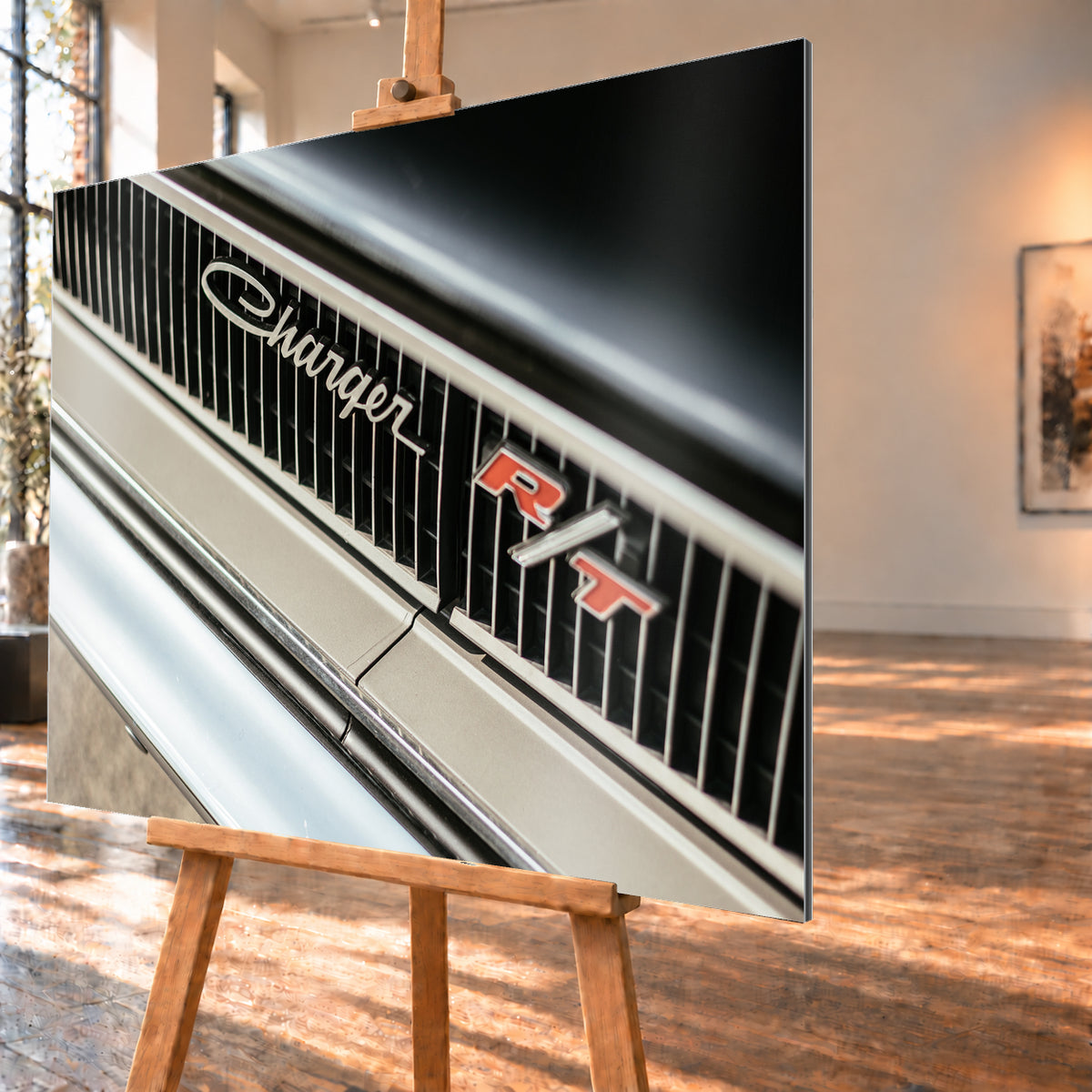 Diagonale Nahaufnahme eines hell lackierten Charger R/T mit schwarzem Lamellengrill, verchromtem Charger-Schriftzug und rot-weißem R/T-Emblem; Chromleisten reflektieren helles Licht; Visualisierung als Alu gebürstet-Wandbild, schräg von der Seite auf einer Holzstaffelei.