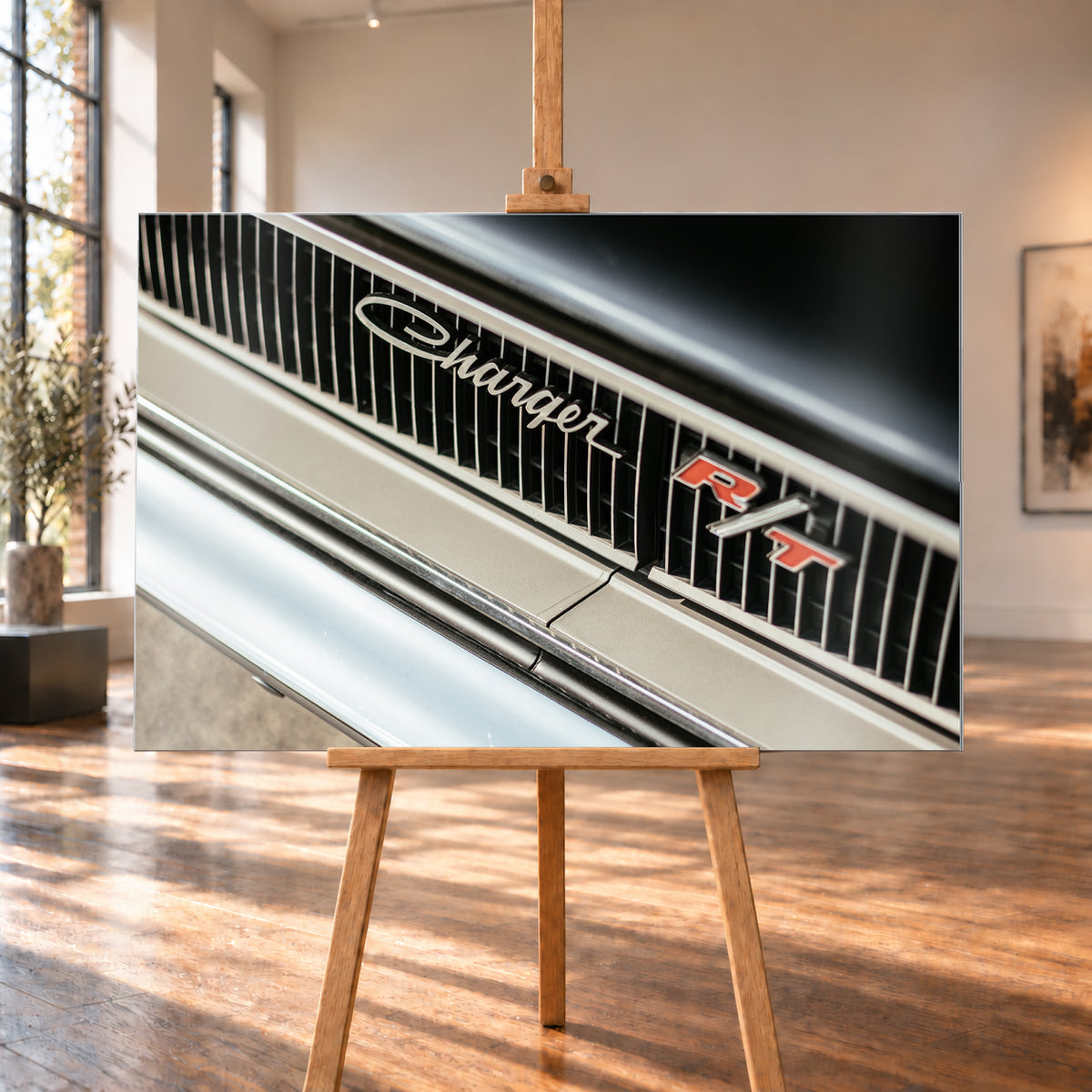 Diagonale Nahaufnahme eines hell lackierten Charger R/T mit schwarzem Lamellengrill, verchromtem Charger-Schriftzug und rot-weißem R/T-Emblem; Chromleisten reflektieren helles Licht; Visualisierung als Alu gebürstet-Wandbild, auf einer Holzstaffelei in einem hellen Galerieraum.