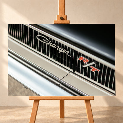 Diagonale Nahaufnahme eines hell lackierten Charger R/T mit schwarzem Lamellengrill, verchromtem Charger-Schriftzug und rot-weißem R/T-Emblem; Chromleisten reflektieren helles Licht; Visualisierung als Leinwand-Wandbild, frontal auf einer Holzstaffelei.