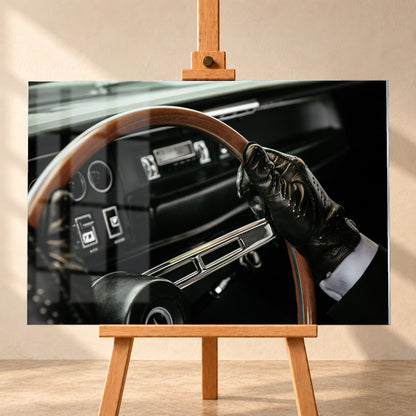 Nahaufnahme eines dunklen Fahrzeugcockpits: Eine Hand in schwarzem, perforiertem Handschuh greift ein braunes, holzfarbenes Lenkrad. Rundinstrumente, Radio und Armaturenbrett liegen unscharf im Hintergrund; seitliches Licht hebt Handschuh, Lenkradkranz und metallisch glänzende Speiche hervor; Visualisierung als Acrylglas-Wandbild, frontal auf einer Holzstaffelei.