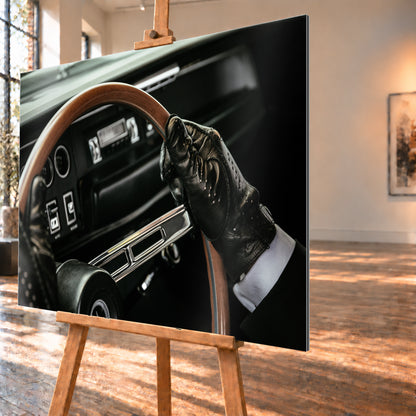 Nahaufnahme eines dunklen Fahrzeugcockpits: Eine Hand in schwarzem, perforiertem Handschuh greift ein braunes, holzfarbenes Lenkrad. Rundinstrumente, Radio und Armaturenbrett liegen unscharf im Hintergrund; seitliches Licht hebt Handschuh, Lenkradkranz und metallisch glänzende Speiche hervor; Visualisierung als Alu gebürstet-Wandbild, schräg von der Seite auf einer Holzstaffelei.