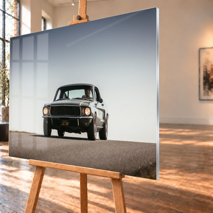 Dunkelgrüner klassischer Ford Mustang in tiefer Frontansicht auf Asphalt, mit eingeschalteten Rundscheinwerfern, sichtbarem Fahrer und weitem hellgrauem Himmel; Visualisierung als Acrylglas-Wandbild, schräg von der Seite auf einer Holzstaffelei.