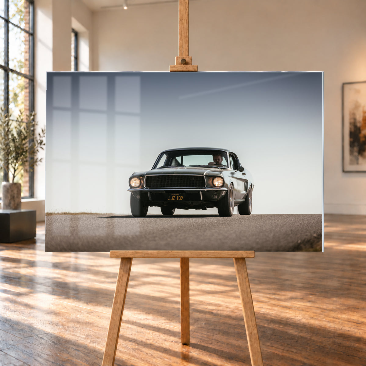 Dunkelgrüner klassischer Ford Mustang in tiefer Frontansicht auf Asphalt, mit eingeschalteten Rundscheinwerfern, sichtbarem Fahrer und weitem hellgrauem Himmel; Visualisierung als Acrylglas-Wandbild, auf einer Holzstaffelei in einem hellen Galerieraum.