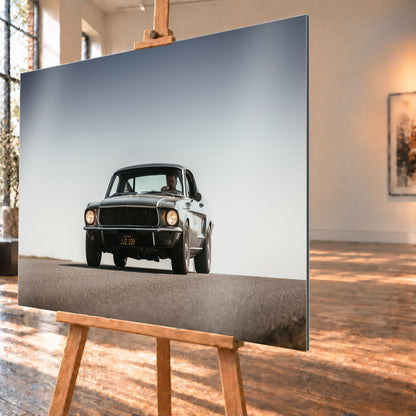 Dunkelgrüner klassischer Ford Mustang in tiefer Frontansicht auf Asphalt, mit eingeschalteten Rundscheinwerfern, sichtbarem Fahrer und weitem hellgrauem Himmel; Visualisierung als Alu gebürstet-Wandbild, schräg von der Seite auf einer Holzstaffelei.