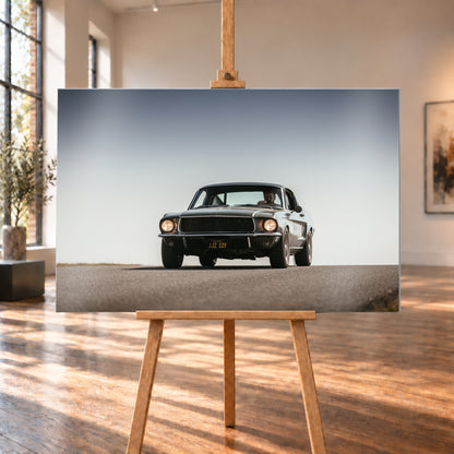 Dunkelgrüner klassischer Ford Mustang in tiefer Frontansicht auf Asphalt, mit eingeschalteten Rundscheinwerfern, sichtbarem Fahrer und weitem hellgrauem Himmel; Visualisierung als Alu gebürstet-Wandbild, auf einer Holzstaffelei in einem hellen Galerieraum.