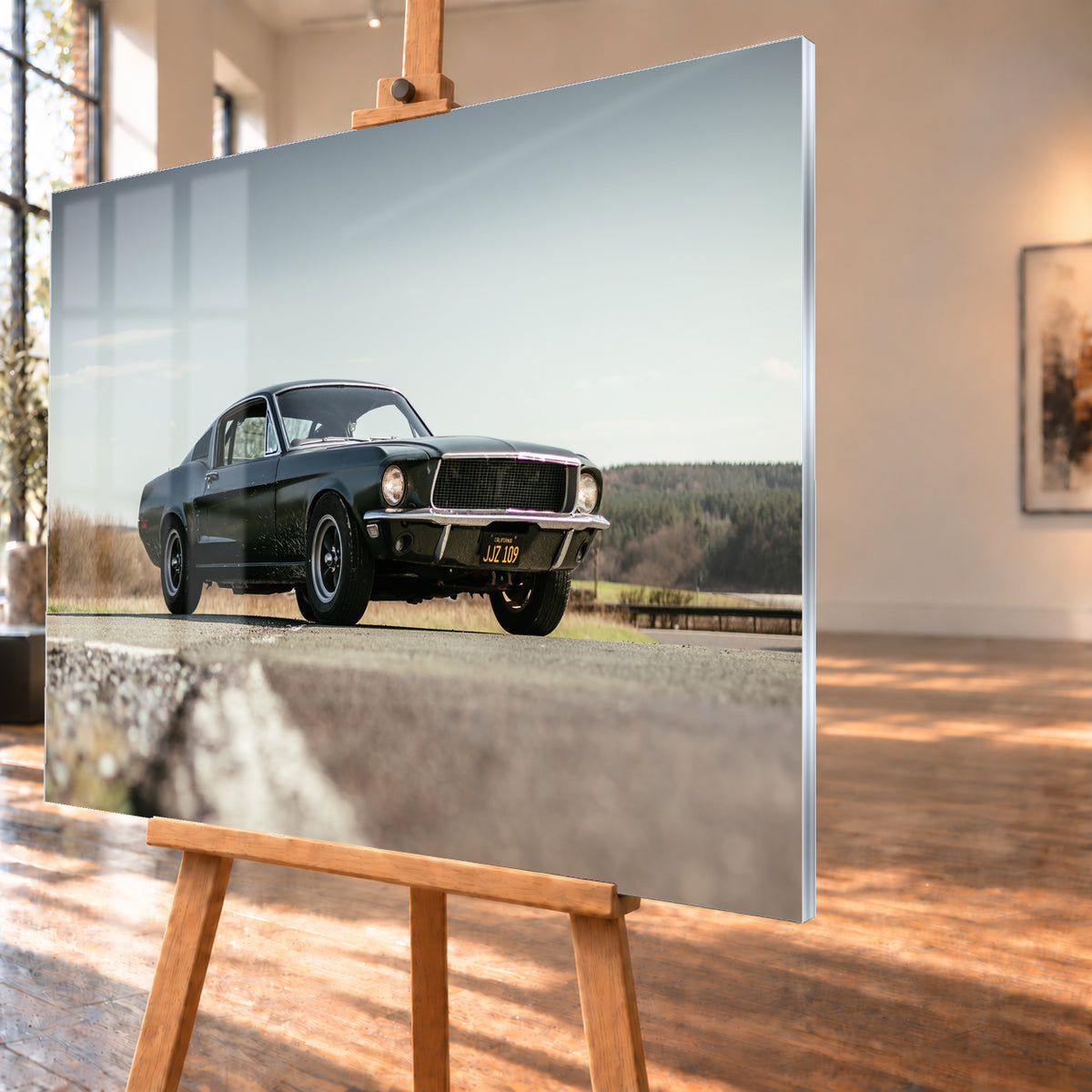 Dunkelgrüner Ford Mustang in niedriger Dreiviertelansicht von vorn auf Asphalt; dahinter offene Felder, ein bewaldeter Horizont und heller Himmel bei Tageslicht; Visualisierung als Acrylglas-Wandbild, schräg von der Seite auf einer Holzstaffelei.