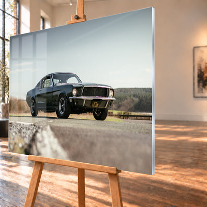 Dunkelgrüner Ford Mustang in niedriger Dreiviertelansicht von vorn auf Asphalt; dahinter offene Felder, ein bewaldeter Horizont und heller Himmel bei Tageslicht; Visualisierung als Acrylglas-Wandbild, schräg von der Seite auf einer Holzstaffelei.