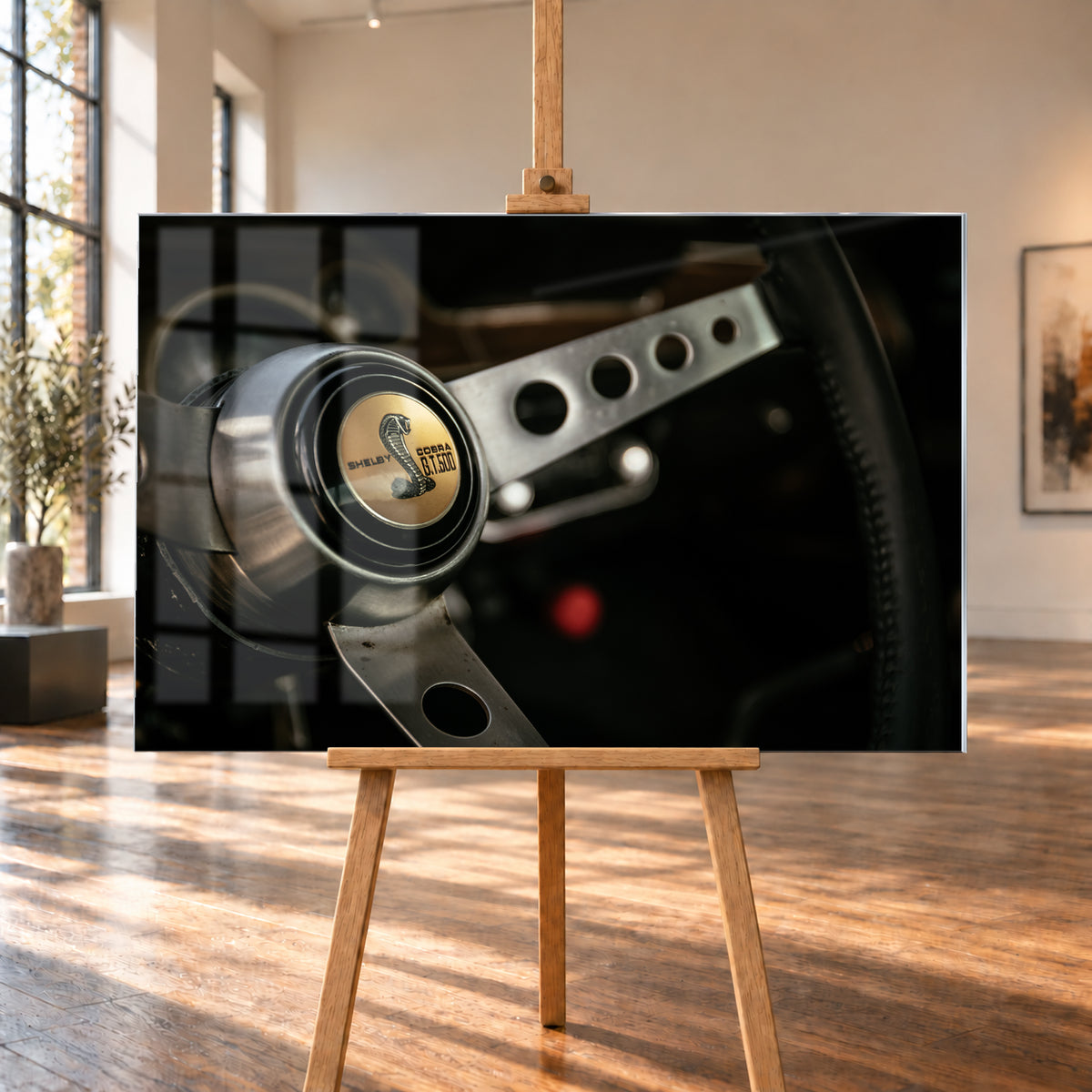 Nahaufnahme eines dreispeichigen Lenkrads mit gelochten silberfarbenen Speichen, schwarzem Kranz und goldenem Shelby-Cobra-GT500-Emblem in einem dunklen, warm beleuchteten Cockpit; Visualisierung als Acrylglas-Wandbild, auf einer Holzstaffelei in einem hellen Galerieraum.