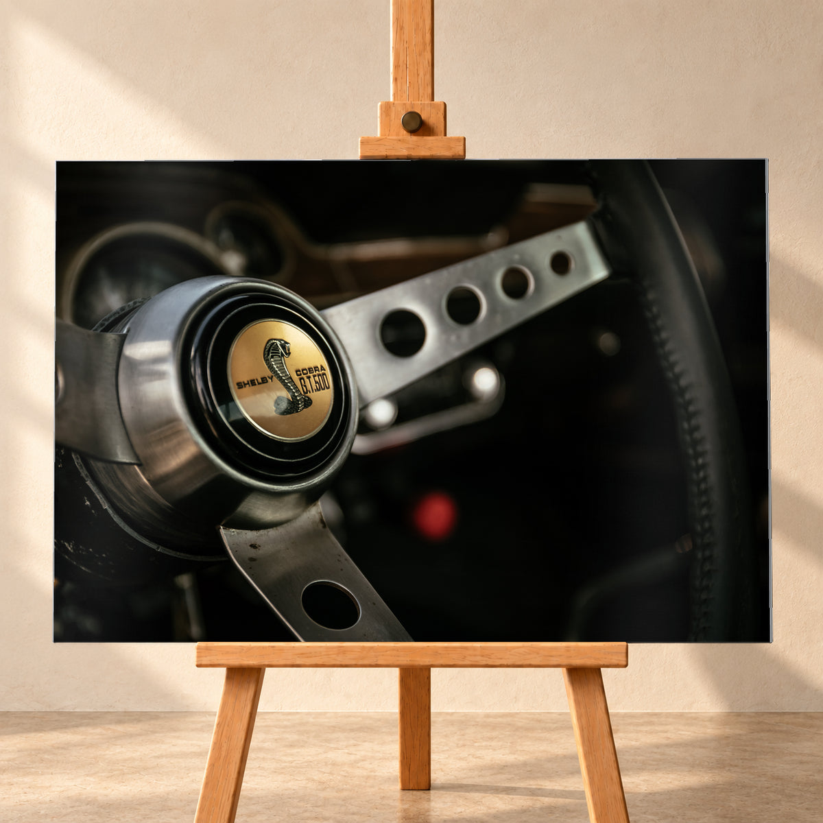 Nahaufnahme eines dreispeichigen Lenkrads mit gelochten silberfarbenen Speichen, schwarzem Kranz und goldenem Shelby-Cobra-GT500-Emblem in einem dunklen, warm beleuchteten Cockpit; Visualisierung als Alu gebürstet-Wandbild, frontal auf einer Holzstaffelei.