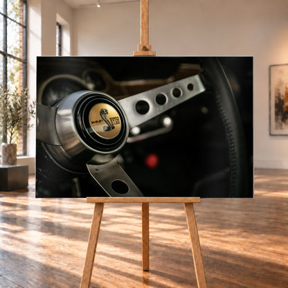 Nahaufnahme eines dreispeichigen Lenkrads mit gelochten silberfarbenen Speichen, schwarzem Kranz und goldenem Shelby-Cobra-GT500-Emblem in einem dunklen, warm beleuchteten Cockpit; Visualisierung als Alu gebürstet-Wandbild, auf einer Holzstaffelei in einem hellen Galerieraum.
