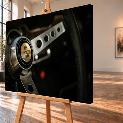 Nahaufnahme eines dreispeichigen Lenkrads mit gelochten silberfarbenen Speichen, schwarzem Kranz und goldenem Shelby-Cobra-GT500-Emblem in einem dunklen, warm beleuchteten Cockpit; Visualisierung als Leinwand-Wandbild, schräg von der Seite auf einer Holzstaffelei.