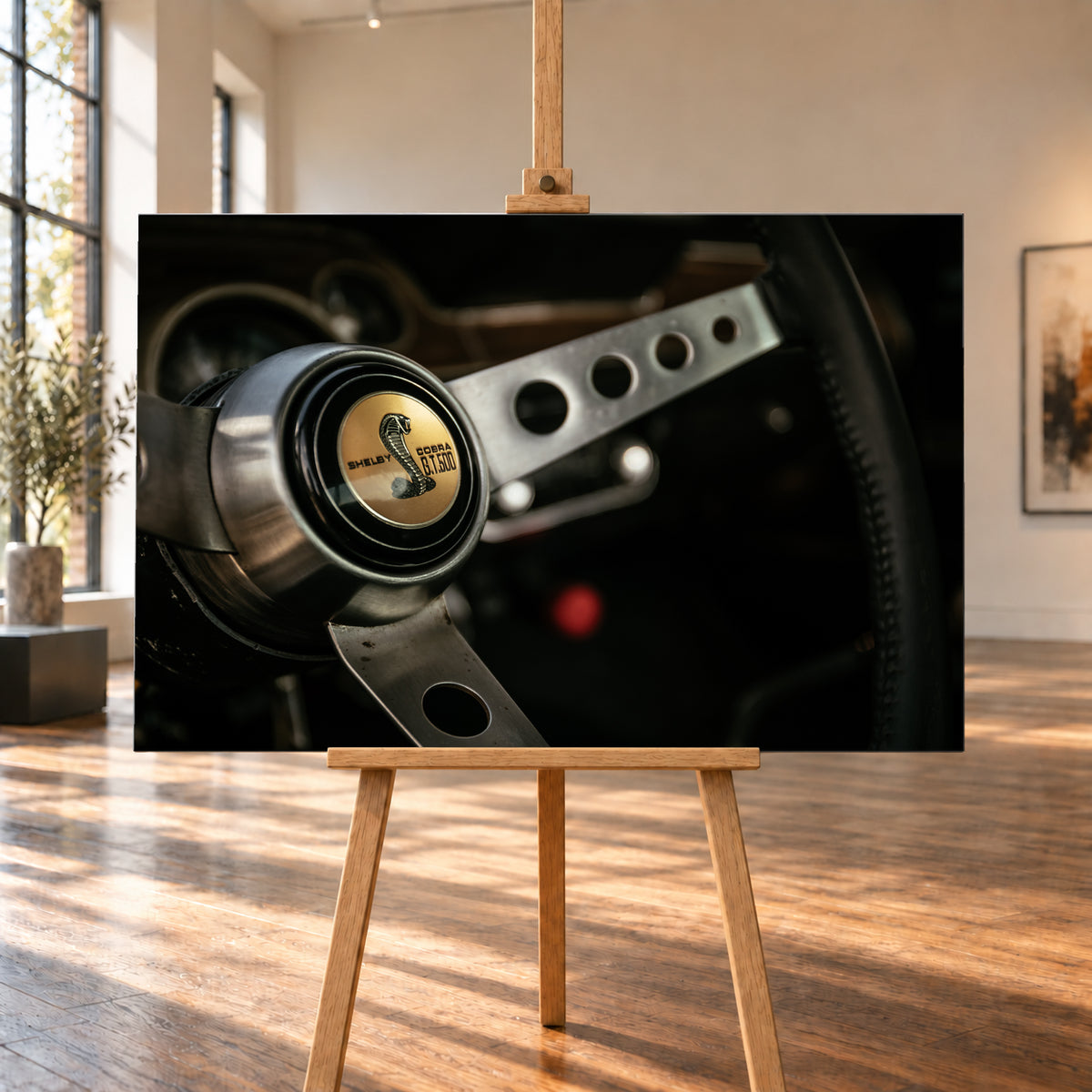 Nahaufnahme eines dreispeichigen Lenkrads mit gelochten silberfarbenen Speichen, schwarzem Kranz und goldenem Shelby-Cobra-GT500-Emblem in einem dunklen, warm beleuchteten Cockpit; Visualisierung als Leinwand-Wandbild, auf einer Holzstaffelei in einem hellen Galerieraum.