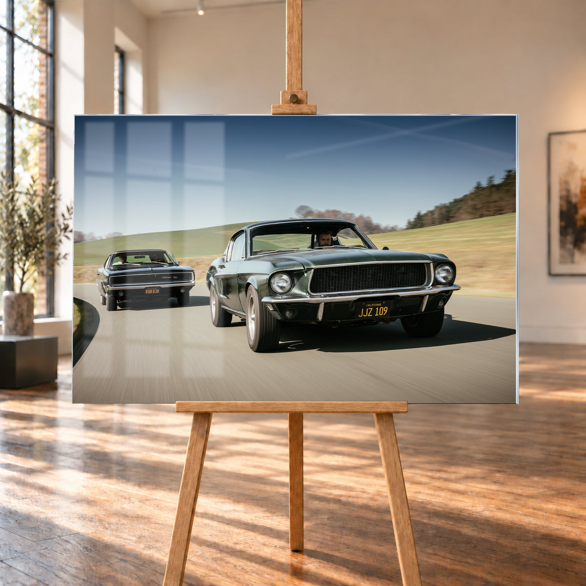 Dynamische Fahraufnahme zweier klassischer Coupés auf einer kurvigen Landstraße. Im Vordergrund fährt ein dunkelgrünes Coupé in Front-Dreiviertelansicht; links dahinter folgt der dunkle Bullitt Charger. Grüne Hügel, Bäume und blauer Himmel rahmen die helle Szene, während verwischte Straße und Umgebung Bewegung vermitteln; Visualisierung als Acrylglas-Wandbild, auf einer Holzstaffelei in einem hellen Galerieraum.