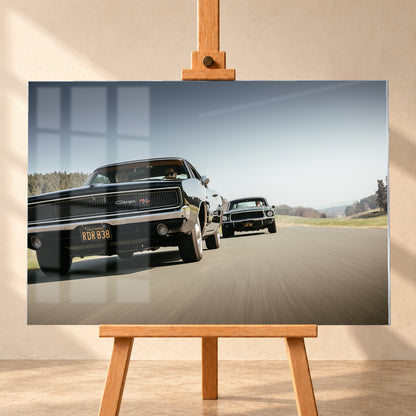 Schwarzer Dodge Charger R/T aus tiefer Frontperspektive auf einer Landstraße; dahinter folgt ein zweites dunkles klassisches Coupé. Heller Himmel, Felder und Bäume umgeben die Straße bei Tageslicht; Visualisierung als Acrylglas-Wandbild, frontal auf einer Holzstaffelei.