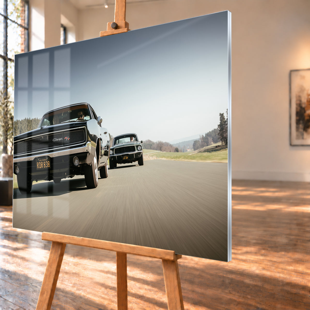 Schwarzer Dodge Charger R/T aus tiefer Frontperspektive auf einer Landstraße; dahinter folgt ein zweites dunkles klassisches Coupé. Heller Himmel, Felder und Bäume umgeben die Straße bei Tageslicht; Visualisierung als Acrylglas-Wandbild, schräg von der Seite auf einer Holzstaffelei.