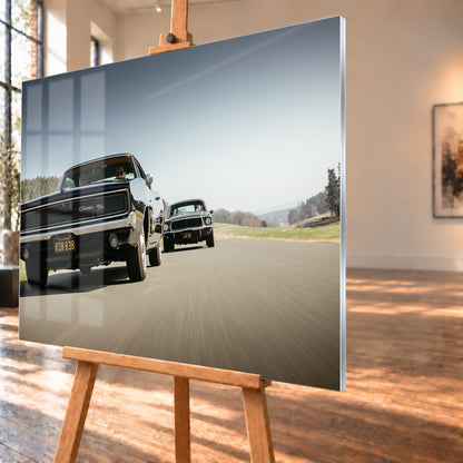 Schwarzer Dodge Charger R/T aus tiefer Frontperspektive auf einer Landstraße; dahinter folgt ein zweites dunkles klassisches Coupé. Heller Himmel, Felder und Bäume umgeben die Straße bei Tageslicht; Visualisierung als Acrylglas-Wandbild, schräg von der Seite auf einer Holzstaffelei.