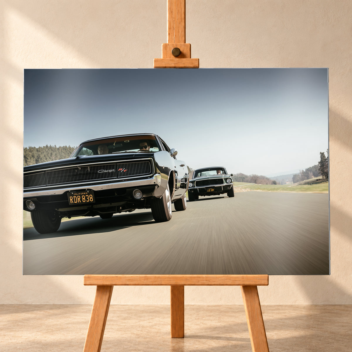 Schwarzer Dodge Charger R/T aus tiefer Frontperspektive auf einer Landstraße; dahinter folgt ein zweites dunkles klassisches Coupé. Heller Himmel, Felder und Bäume umgeben die Straße bei Tageslicht; Visualisierung als Alu gebürstet-Wandbild, frontal auf einer Holzstaffelei.