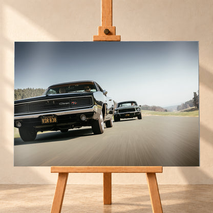 Schwarzer Dodge Charger R/T aus tiefer Frontperspektive auf einer Landstraße; dahinter folgt ein zweites dunkles klassisches Coupé. Heller Himmel, Felder und Bäume umgeben die Straße bei Tageslicht; Visualisierung als Alu gebürstet-Wandbild, frontal auf einer Holzstaffelei.