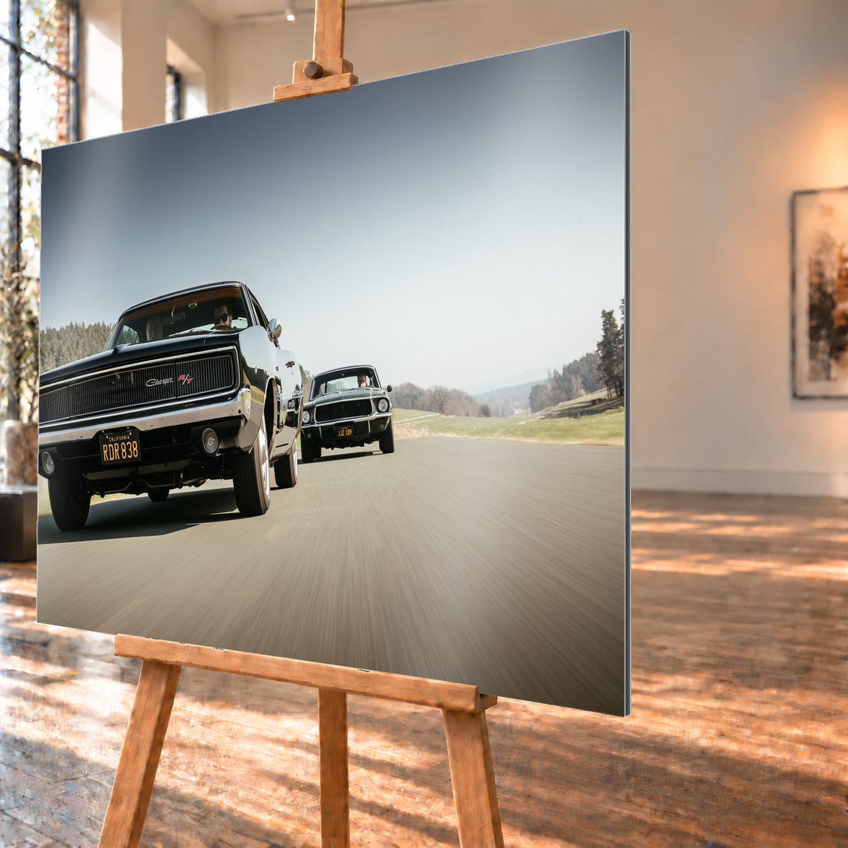 Schwarzer Dodge Charger R/T aus tiefer Frontperspektive auf einer Landstraße; dahinter folgt ein zweites dunkles klassisches Coupé. Heller Himmel, Felder und Bäume umgeben die Straße bei Tageslicht; Visualisierung als Alu gebürstet-Wandbild, schräg von der Seite auf einer Holzstaffelei.