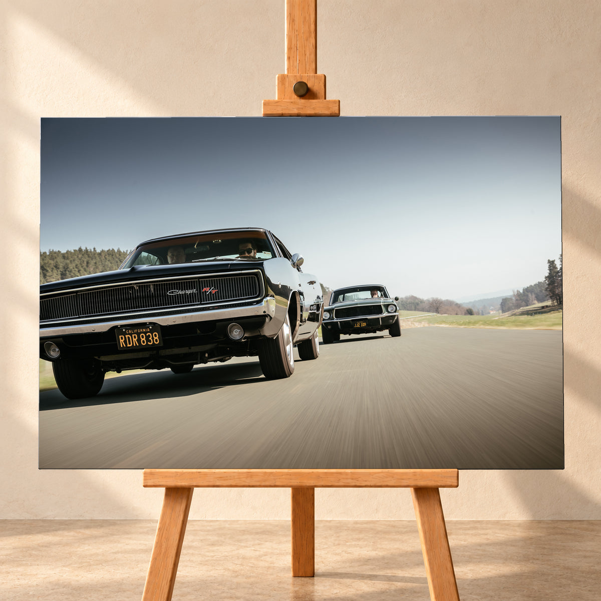 Schwarzer Dodge Charger R/T aus tiefer Frontperspektive auf einer Landstraße; dahinter folgt ein zweites dunkles klassisches Coupé. Heller Himmel, Felder und Bäume umgeben die Straße bei Tageslicht; Visualisierung als Leinwand-Wandbild, frontal auf einer Holzstaffelei.