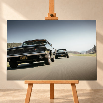 Schwarzer Dodge Charger R/T aus tiefer Frontperspektive auf einer Landstraße; dahinter folgt ein zweites dunkles klassisches Coupé. Heller Himmel, Felder und Bäume umgeben die Straße bei Tageslicht; Visualisierung als Leinwand-Wandbild, frontal auf einer Holzstaffelei.
