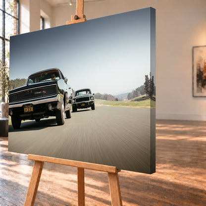 Schwarzer Dodge Charger R/T aus tiefer Frontperspektive auf einer Landstraße; dahinter folgt ein zweites dunkles klassisches Coupé. Heller Himmel, Felder und Bäume umgeben die Straße bei Tageslicht; Visualisierung als Leinwand-Wandbild, schräg von der Seite auf einer Holzstaffelei.