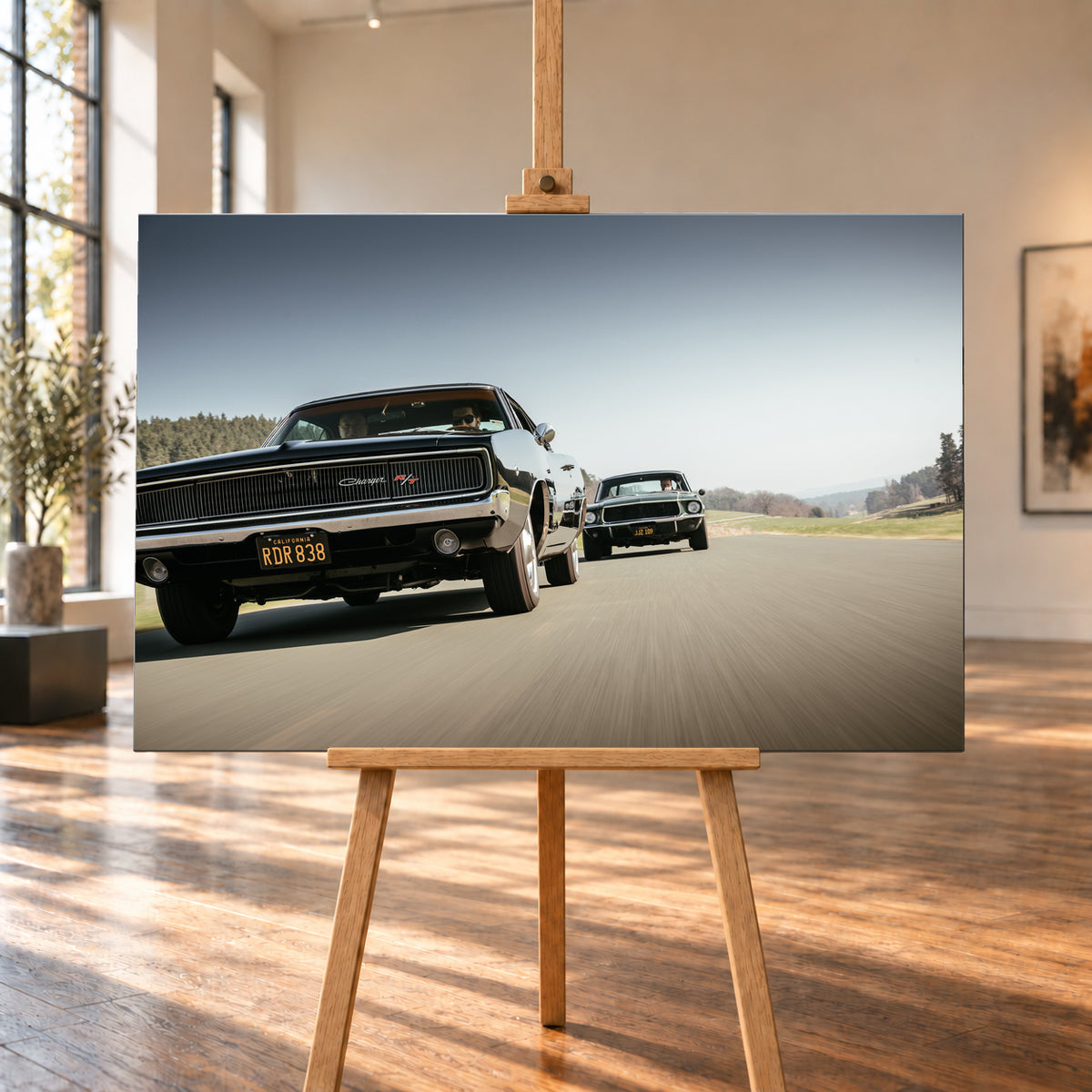Schwarzer Dodge Charger R/T aus tiefer Frontperspektive auf einer Landstraße; dahinter folgt ein zweites dunkles klassisches Coupé. Heller Himmel, Felder und Bäume umgeben die Straße bei Tageslicht; Visualisierung als Leinwand-Wandbild, auf einer Holzstaffelei in einem hellen Galerieraum.