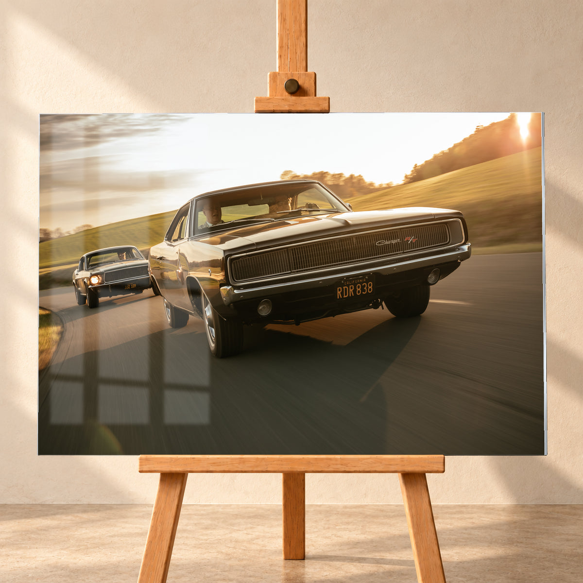 Dunkel erscheinender Dodge Charger R/T aus tiefer Frontperspektive auf einer kurvigen Landstraße; dahinter folgt ein weiteres dunkles Coupé. Goldenes Gegenlicht, grüne Hügel und starke Bewegungsunschärfe prägen die Fahrszene; Visualisierung als Acrylglas-Wandbild, frontal auf einer Holzstaffelei.