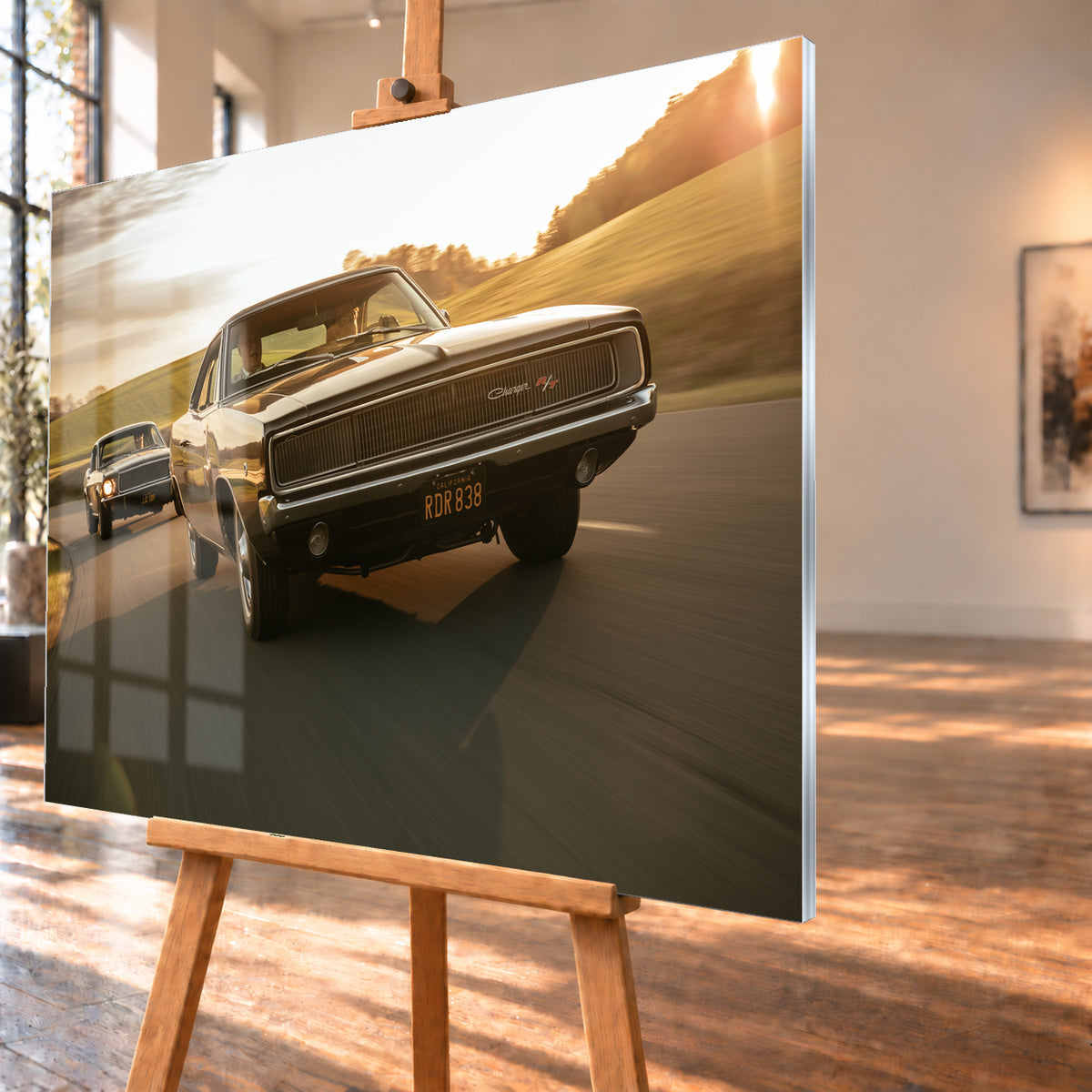 Dunkel erscheinender Dodge Charger R/T aus tiefer Frontperspektive auf einer kurvigen Landstraße; dahinter folgt ein weiteres dunkles Coupé. Goldenes Gegenlicht, grüne Hügel und starke Bewegungsunschärfe prägen die Fahrszene; Visualisierung als Acrylglas-Wandbild, schräg von der Seite auf einer Holzstaffelei.