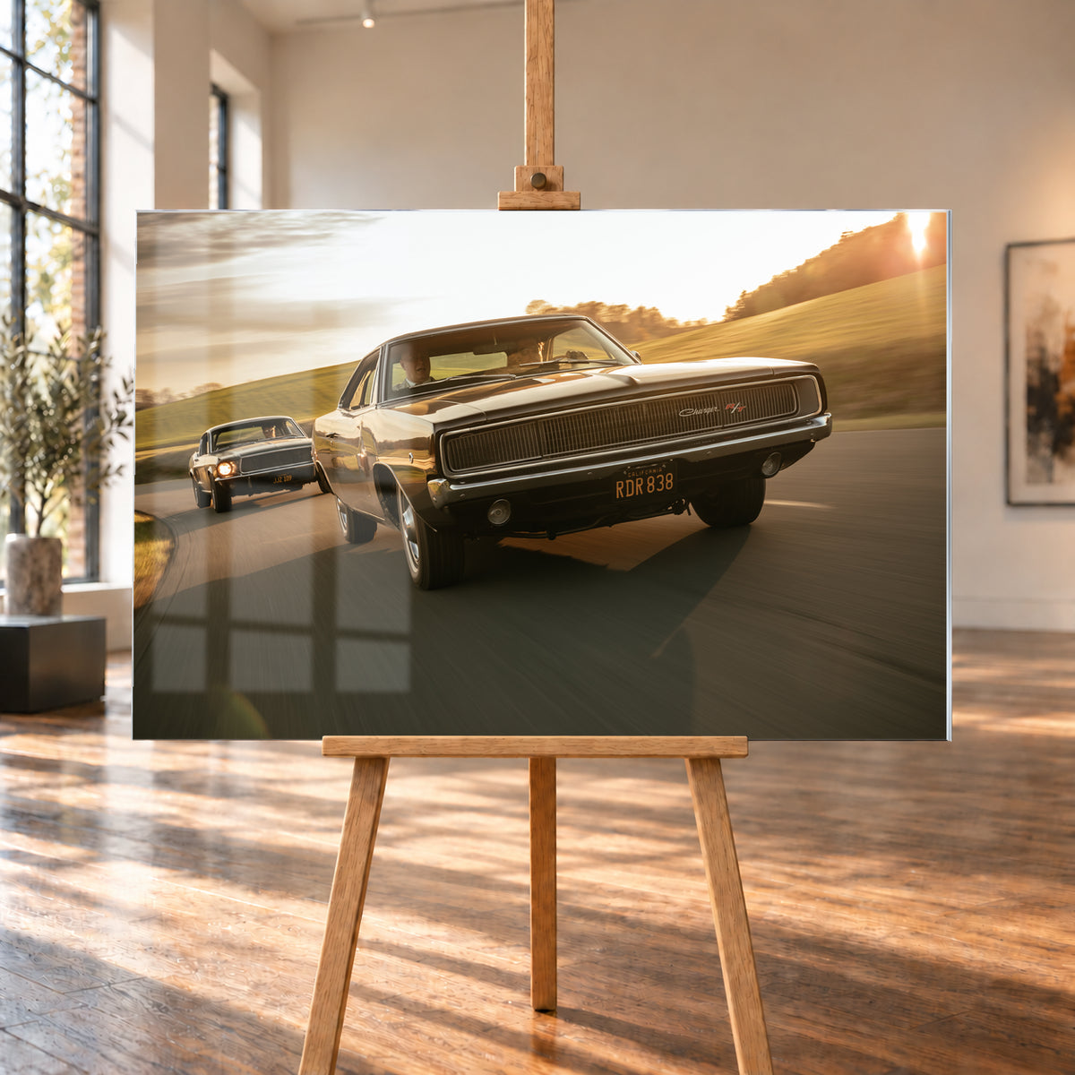 Dunkel erscheinender Dodge Charger R/T aus tiefer Frontperspektive auf einer kurvigen Landstraße; dahinter folgt ein weiteres dunkles Coupé. Goldenes Gegenlicht, grüne Hügel und starke Bewegungsunschärfe prägen die Fahrszene; Visualisierung als Acrylglas-Wandbild, auf einer Holzstaffelei in einem hellen Galerieraum.