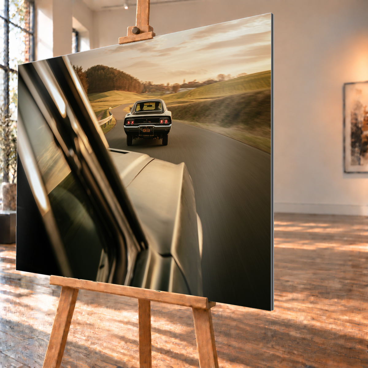 Dunkler Dodge Charger 440 R/T von hinten auf einer kurvigen Landstraße im goldenen Abendlicht, fotografiert aus einem zweiten fahrenden Auto; rote Rückleuchten, Wiesen und Bewegungsunschärfe; Visualisierung als Alu gebürstet-Wandbild, schräg von der Seite auf einer Holzstaffelei.