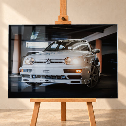 Weißer VW Jetta in niedriger Front-Dreiviertelansicht auf einem dunklen Parkdeck, mit eingeschalteten Scheinwerfern, Dekoren und Startnummer 4. Orangefarbene Säulen, Lichtreflexe und ein unscharfes dunkles Fahrzeug im Hintergrund rahmen die Szene; Visualisierung als Acrylglas-Wandbild, frontal auf einer Holzstaffelei.