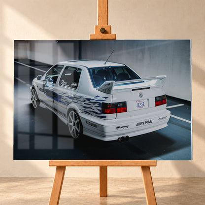 Weißer VW Jetta in rückwärtiger Dreiviertelansicht in einem Parkbereich, mit blauen Flammengrafiken, großem Heckflügel, hellen Mehrspeichenrädern und roten Rückleuchten bei kühlem Licht; Visualisierung als Acrylglas-Wandbild, frontal auf einer Holzstaffelei.