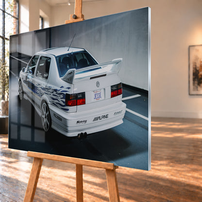 Weißer VW Jetta in rückwärtiger Dreiviertelansicht in einem Parkbereich, mit blauen Flammengrafiken, großem Heckflügel, hellen Mehrspeichenrädern und roten Rückleuchten bei kühlem Licht; Visualisierung als Acrylglas-Wandbild, schräg von der Seite auf einer Holzstaffelei.