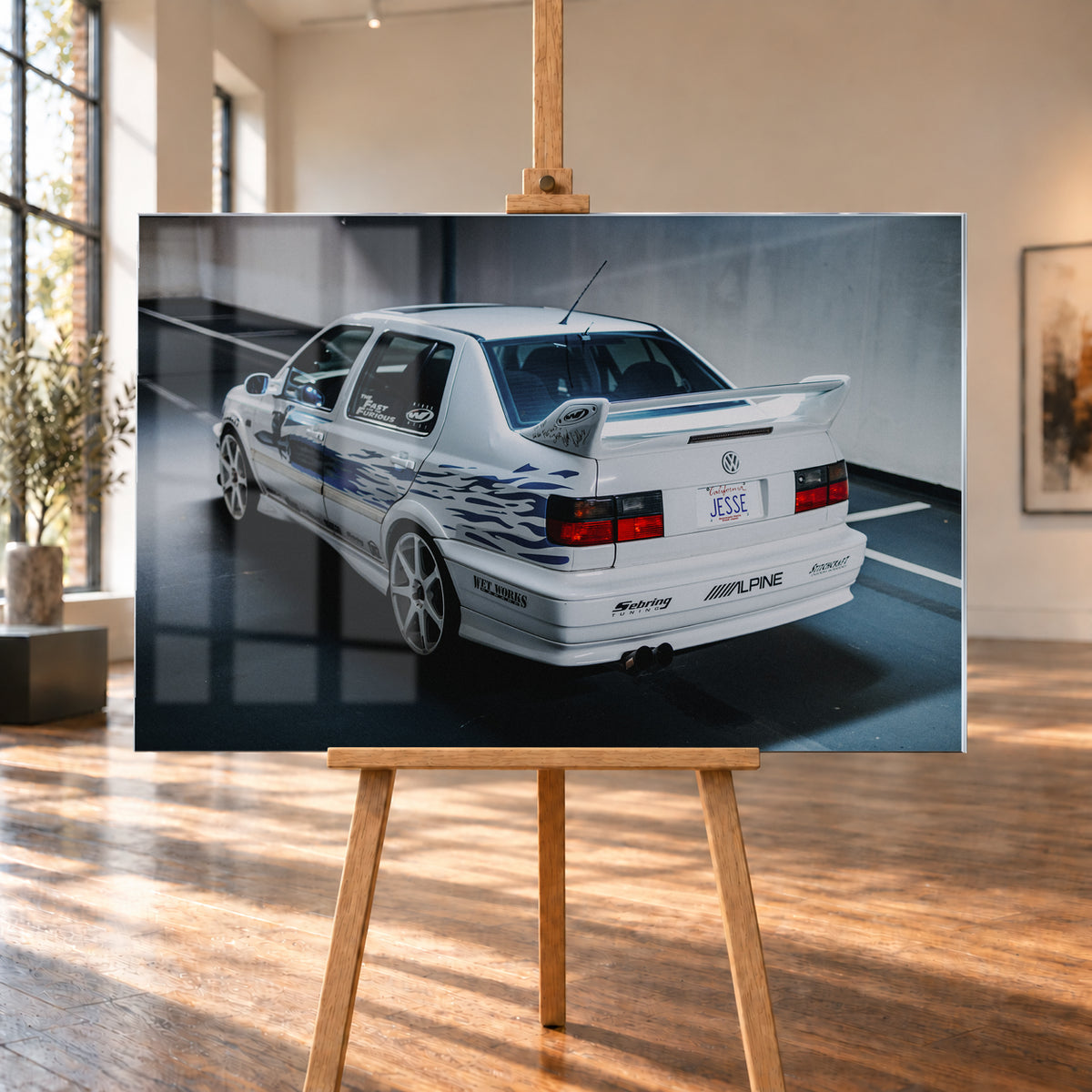 Weißer VW Jetta in rückwärtiger Dreiviertelansicht in einem Parkbereich, mit blauen Flammengrafiken, großem Heckflügel, hellen Mehrspeichenrädern und roten Rückleuchten bei kühlem Licht; Visualisierung als Acrylglas-Wandbild, auf einer Holzstaffelei in einem hellen Galerieraum.