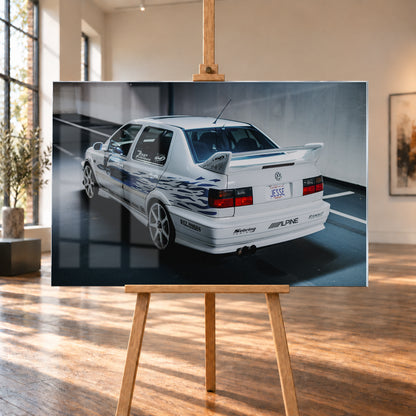 Weißer VW Jetta in rückwärtiger Dreiviertelansicht in einem Parkbereich, mit blauen Flammengrafiken, großem Heckflügel, hellen Mehrspeichenrädern und roten Rückleuchten bei kühlem Licht; Visualisierung als Acrylglas-Wandbild, auf einer Holzstaffelei in einem hellen Galerieraum.
