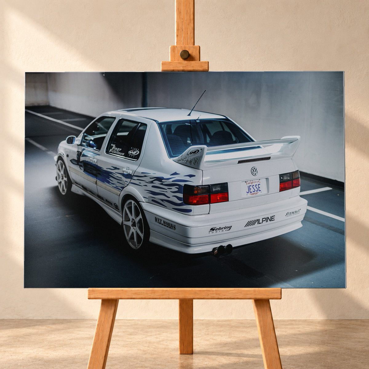 Weißer VW Jetta in rückwärtiger Dreiviertelansicht in einem Parkbereich, mit blauen Flammengrafiken, großem Heckflügel, hellen Mehrspeichenrädern und roten Rückleuchten bei kühlem Licht; Visualisierung als Alu gebürstet-Wandbild, frontal auf einer Holzstaffelei.