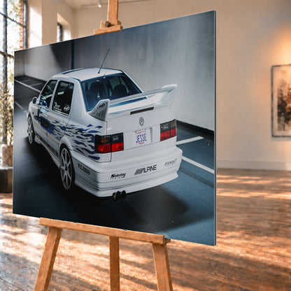 Weißer VW Jetta in rückwärtiger Dreiviertelansicht in einem Parkbereich, mit blauen Flammengrafiken, großem Heckflügel, hellen Mehrspeichenrädern und roten Rückleuchten bei kühlem Licht; Visualisierung als Alu gebürstet-Wandbild, schräg von der Seite auf einer Holzstaffelei.