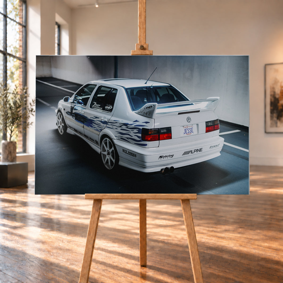 Weißer VW Jetta in rückwärtiger Dreiviertelansicht in einem Parkbereich, mit blauen Flammengrafiken, großem Heckflügel, hellen Mehrspeichenrädern und roten Rückleuchten bei kühlem Licht; Visualisierung als Alu gebürstet-Wandbild, auf einer Holzstaffelei in einem hellen Galerieraum.